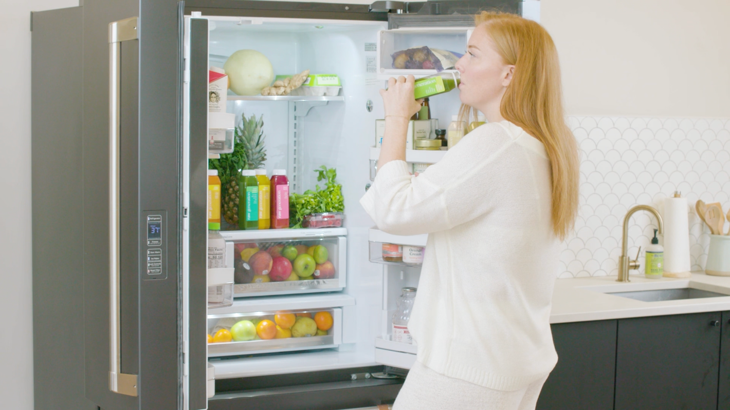 Woman drinking Raw Generation juice in front of a fridge with healthy produce choices
